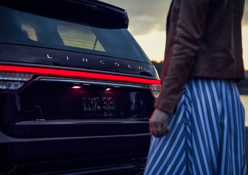 A person is shown near the rear of a 2024 Lincoln Aviator® SUV as the Lincoln Embrace illuminates the rear lights | Hooks Lincoln in Fort Worth TX